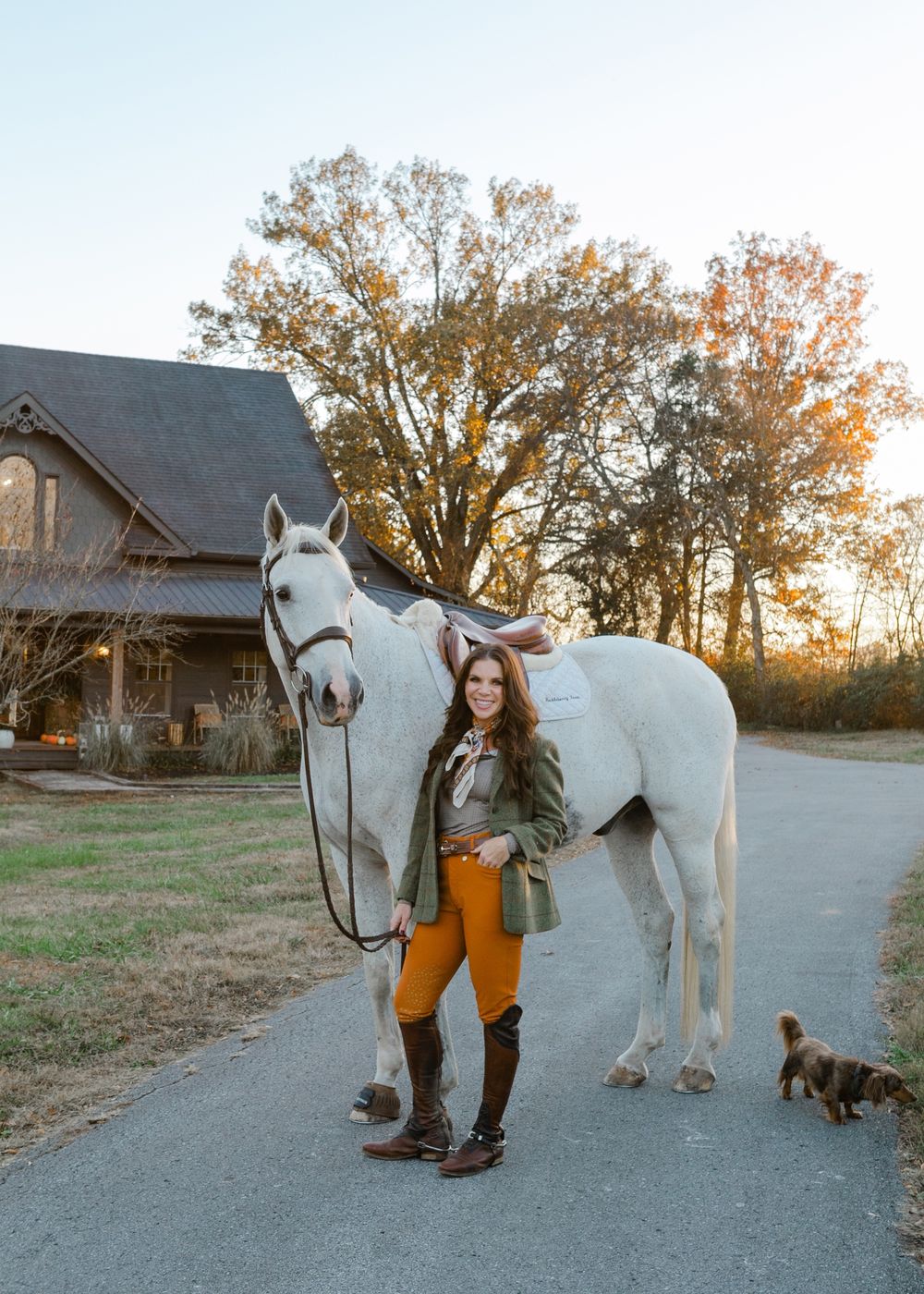 Tiffany standing beside a saddled white horse in front of her farmhouse at sunset, with her dachshund in the lane.