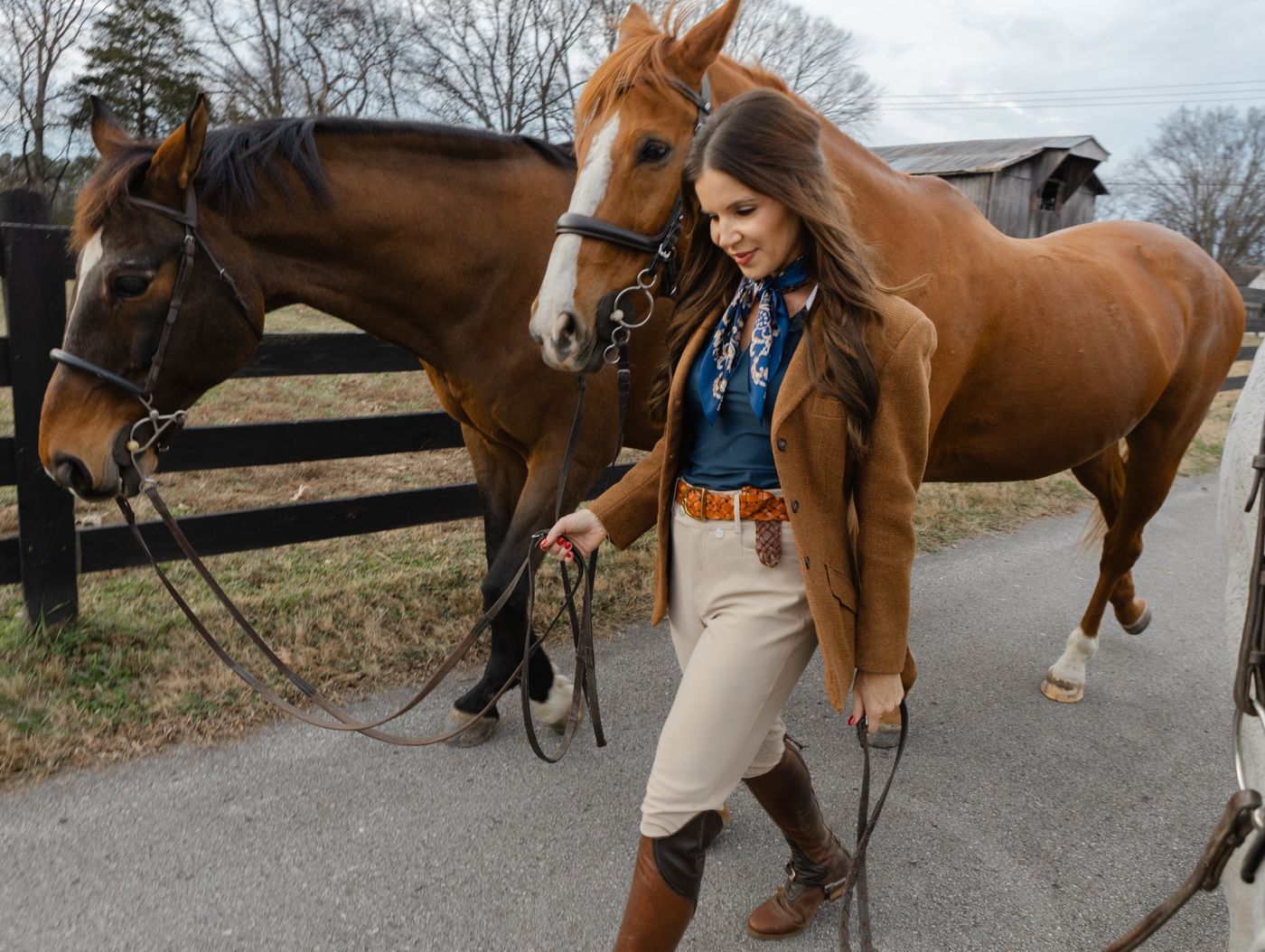 Tiffany leading two horses down a farm lane beside a black board fence, with an old barn behind them.
